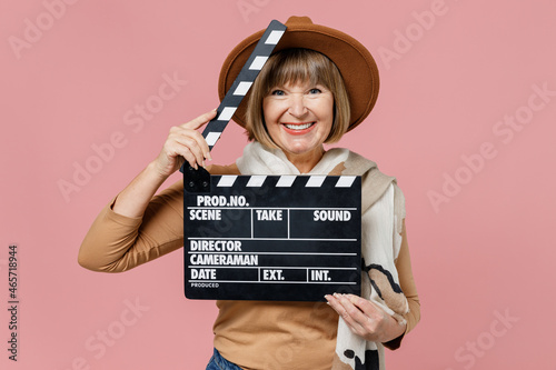 Traveler tourist mature elderly senior lady woman 55 years old wear brown shirt hat scarf holding classic black film making clapperboard isolated on plain pastel light pink background studio portrait