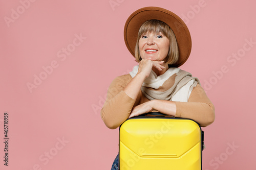 Traveler tourist fun mature elderly senior lady woman 55 years old wear brown shirt hat scarf hold suitcase bag put hand prop up on chin isolated on plain pastel light pink background studio portrait.