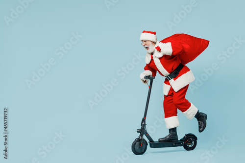 Full body old Santa Claus man 50s in Christmas hat red suit clothes rides kick scooter with gifts bag isolated on plain blue background studio. Happy New Year 2022 celebration merry ho x-mas concept.