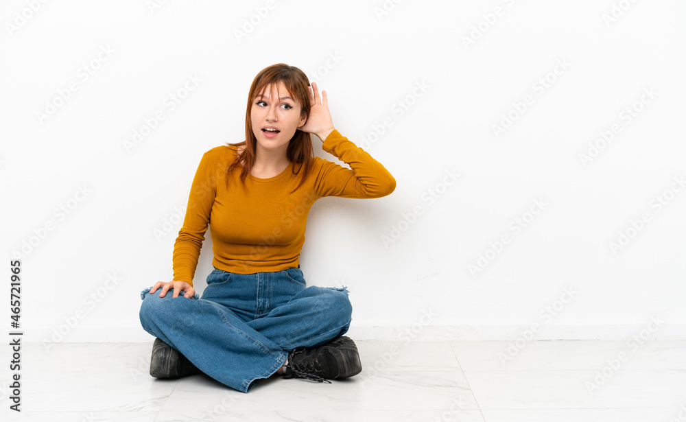 Redhead girl sitting on the floor isolated on white background listening to something by putting hand on the ear Redhead girl sitting on the floor isolated on white background listening to something by putting hand on the ear
