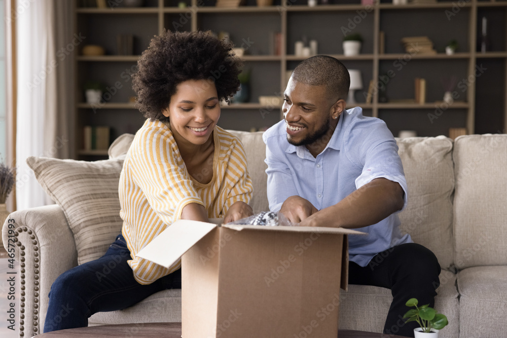 Happy curious young african american family couple opening big carton box, enjoying unpacking internet store order together at home, satisfied with fast delivery service, online shopping concept. Happy curious young african american family couple opening big carton box, enjoying unpacking internet store order together at home, satisfied with fast delivery service, online shopping concept.