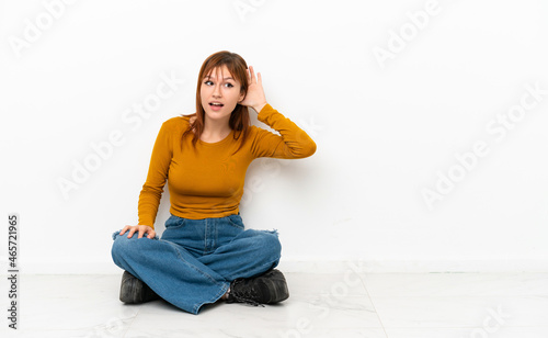 Redhead girl sitting on the floor isolated on white background listening to something by putting hand on the ear