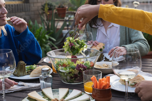 Hand of a woman picking salad from a bowl at a table with vegetarian food next to a group of women on a terrace.