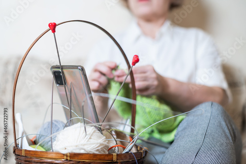 Faceless old woman knits and watches online training on a smartphone. Close-up of female hands with yarn and knitting needles.