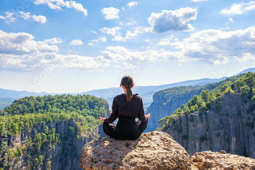 Back view of anonymous peaceful female sitting in lotus pose on edge of rocky cliff above mountains in highland during yoga session photo