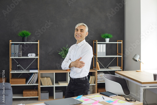 Portrait of happy successful confident self made old businessman at work. Positive senior man in white shirt standing by office desk, looking at camera and smiling. Business concept. Story of success.