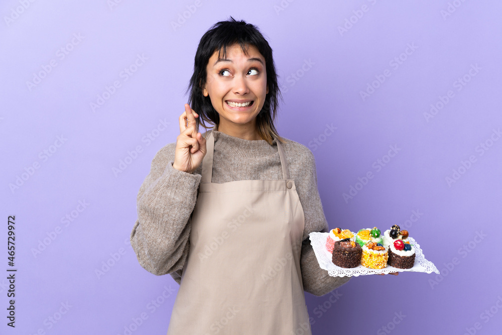 Young Uruguayan woman holding lots of different mini cakes over isolated purple background with fingers crossing Young Uruguayan woman holding lots of different mini cakes over isolated purple background with fingers crossing
