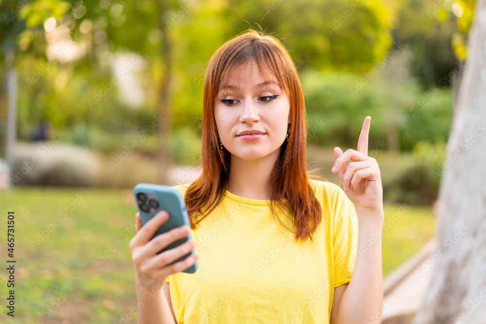 Young pretty redhead woman at outdoors using mobile phone and lifting finger