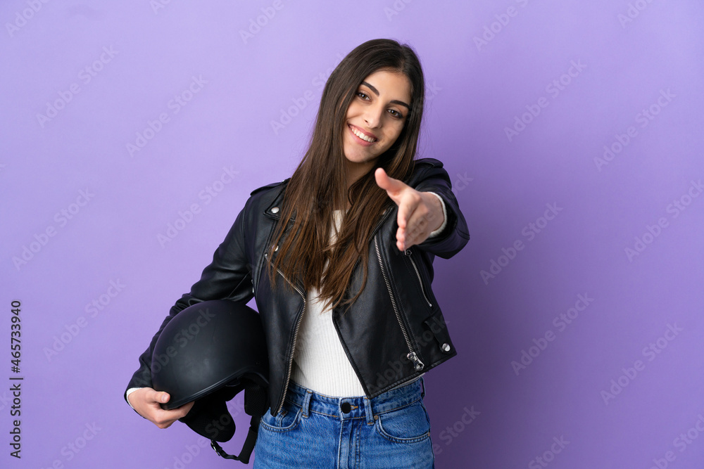Fototapeta premium Young caucasian woman with a motorcycle helmet isolated on purple background shaking hands for closing a good deal