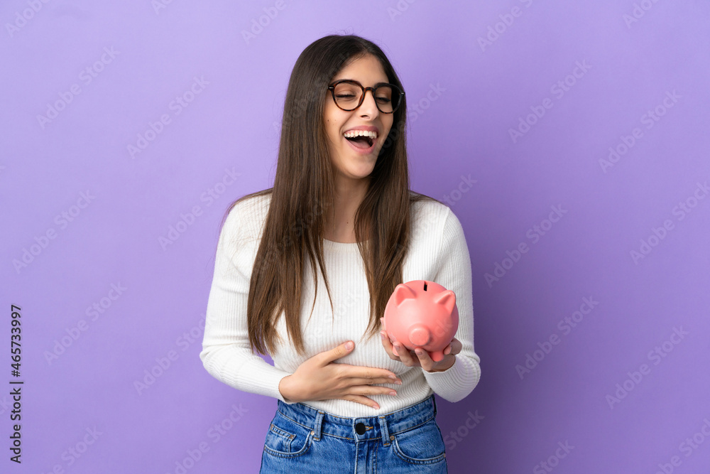 Young caucasian woman holding a piggybank isolated on purple background smiling a lot Young caucasian woman holding a piggybank isolated on purple background smiling a lot
