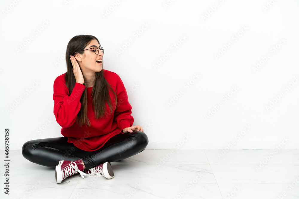 Young woman sitting on the floor isolated on white background listening to something by putting hand on the ear Young woman sitting on the floor isolated on white background listening to something by putting hand on the ear