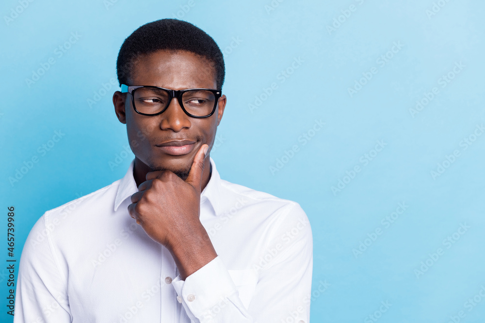 Photo of pretty serious dark skin man wear white shirt arm chin looking empty space isolated blue color background Photo of pretty serious dark skin man wear white shirt arm chin looking empty space isolated blue color background
