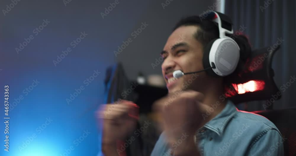 Young Asian man playing online computer video game, colorful lighting broadcast streaming live at home. Ecstatic celebration winning a match. Gamer lifestyle, E-Sport online gaming technology concept