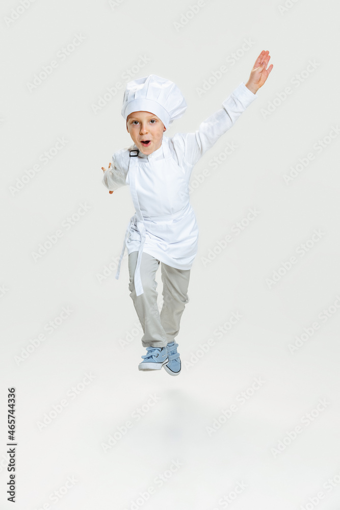 One little preschool boy in white cook uniform and huge chef's hat running isolated on white studio background.