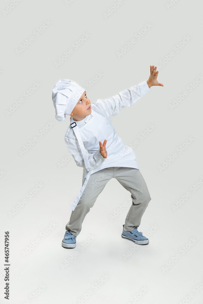 Studio shot of little scared boy in white cook uniform and huge chef's hat standing isolated on white studio background.
