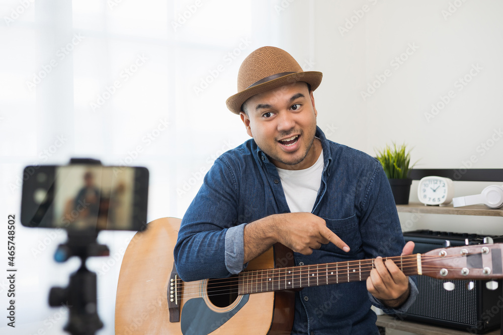 Obraz premium Young asian musician teaching the guitar class online live streaming with smartphone with student. Indian man playing acoustic guitar while singing a song and record by cell phone shaed online social