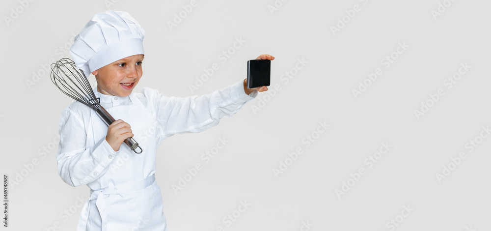 Close-up portrait of little happy boy in white cook uniform and huge chef's hat using phone isolated on white studio background. Close-up portrait of little happy boy in white cook uniform and huge chef's hat using phone isolated on white studio background.