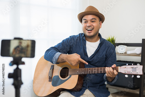 Young asian musician teaching the guitar class online live streaming with smartphone with student. Indian man playing acoustic guitar while singing a song and record by cell phone shaed online social