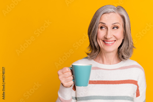 Photo of pretty adorable lady pensioner wear striped pullover drinking looking empty space smiling isolated yellow color background