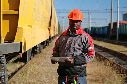 Railway man with tablet computer at freight train terminal. Railroad man in uniform and red hard hat with computer. Railway employee holds in hands tablet pc and look at the camera