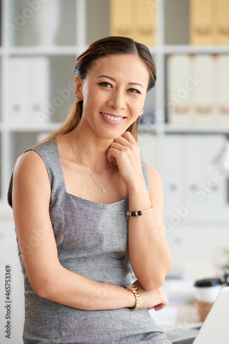 Portrait of smiling young Vietnamese businesswoman in jewelries touching neck against shelves with folders in office Free Download Portrait of smiling young Vietnamese businesswoman in jewelries touching neck against shelves with folders in office - GFXLoad