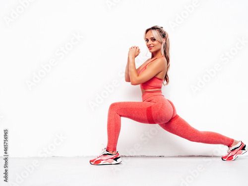 Fototapeta Naklejka Na Ścianę i Meble -  Fitness smiling woman in pink sports clothing. Young beautiful model with perfect body. Female posing in the street near white wall at sunny day. Doing forward lunges. Stretching out before training
