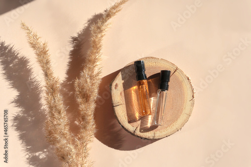 Two glass perfume samples with transparent brown liquid on wooden tray lying on beige background with pampas grass. Luxury and natural cosmetics presentation. Testers on woodcut in sunlight. Top view