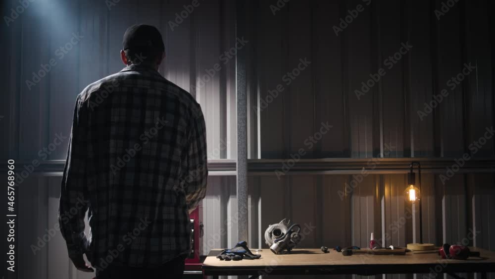 A tall man walking into an industrial garage with a light shining down, opening a red toolbox and taking out tools. On the workbench in the background you can see a turbocharger, gloves, hammer, work 
