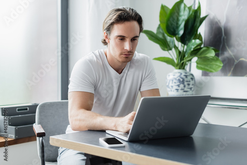 concentrated freelancer looking at laptop near smartphone on desk while working from home