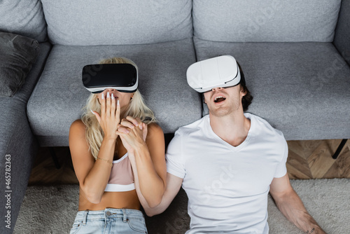 high angle view of shocked couple in vr headsets sitting near grey couch