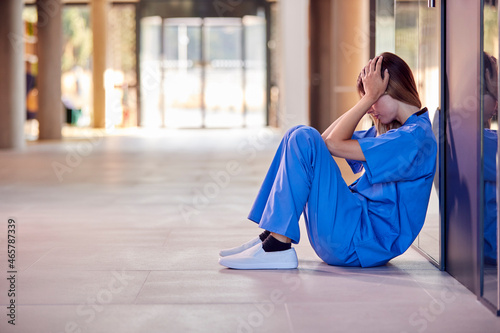 Tired And Overworked Medical Worker In Scrubs Sitting On Floor Of Hospital Corridor During Break