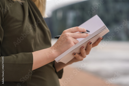 Unrecognizable person in a business park pointing notes in the agenda, young blonde in a green shirt leaning against a wooden wall, copy space and paste