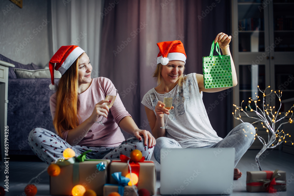 Two cheerful girlfriends open xmas gift boxes during online video conference with family on laptop. Sisters exchange gifts in home interior with festive decorations.
