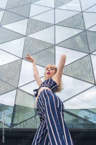 Fashionable posing of a young blonde in a glass building, blonde girl in a blue striped suit, arms up