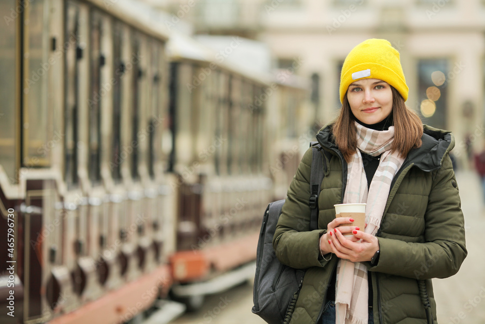 Travel and coffee concept: happy smiling young woman drinking hot beverage at street of European city. Model wearing green coat, white and beige scarf. Copyspace. Outdoor shot Travel and coffee concept: happy smiling young woman drinking hot beverage at street of European city. Model wearing green coat, white and beige scarf. Copyspace. Outdoor shot