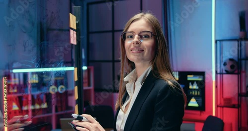 Beautiful positive confident creative brown-haired businesswoman in glasses writing on glass wall importnat notes and then looking into camera with sincerely smile in evening office photo