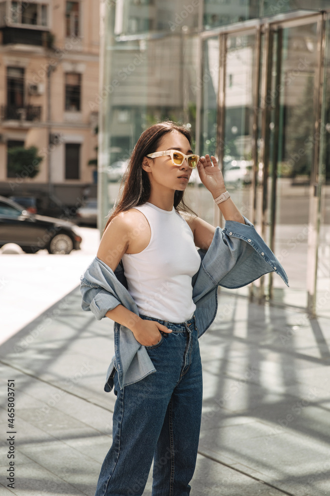 Fototapeta premium Half-length image of serious young woman standing in front of panoramic building. Brunette with glasses confidently looks in front of with her hand in her pocket. Lifestyle concept