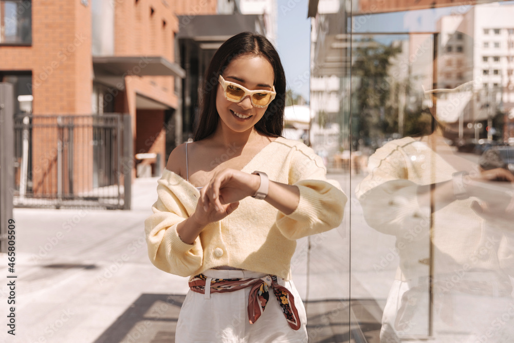 Fashionable asian girl in sunglasses looks at her wristwatch in summer in european city. Cute brunette dressed in light sweater and white jeans. Urban life concept Fashionable asian girl in sunglasses looks at her wristwatch in summer in european city. Cute brunette dressed in light sweater and white jeans. Urban life concept