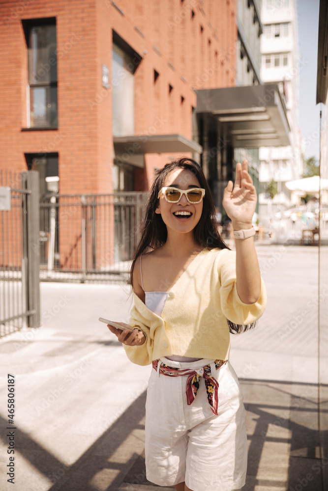 Cute asian woman waving her hand at camera smiling broadly on background of brick house. Brunette with long hair in yellow jacket and white loose shorts with phone in her hands. Cute asian woman waving her hand at camera smiling broadly on background of brick house. Brunette with long hair in yellow jacket and white loose shorts with phone in her hands.