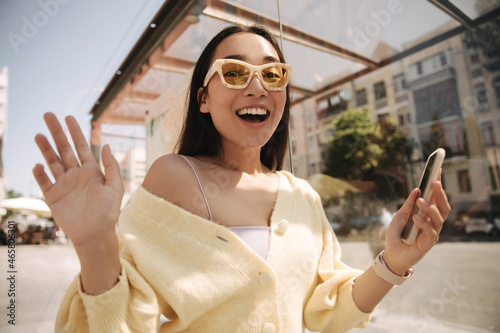 Pleasantly surprised beautiful asian lady with her mouth wide open looks at camera. Brunette in sunglasses and phone waving her hands spends time on street. Summer playful mood concept