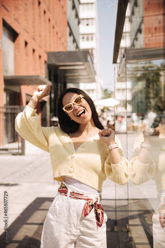 Mischievous, carefree young asian beauty moves in dance with open mouth in street photo. Girl in light yellow sweater with button, white trousers and sunglasses fashionable.