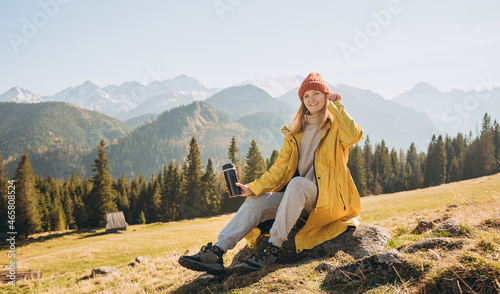 Traveler girl drinking tea from thermos cup over nature background. Mountain peaks. Freedom, happiness, travel and vacations concept, outdoor activities, woman wearing red hat
