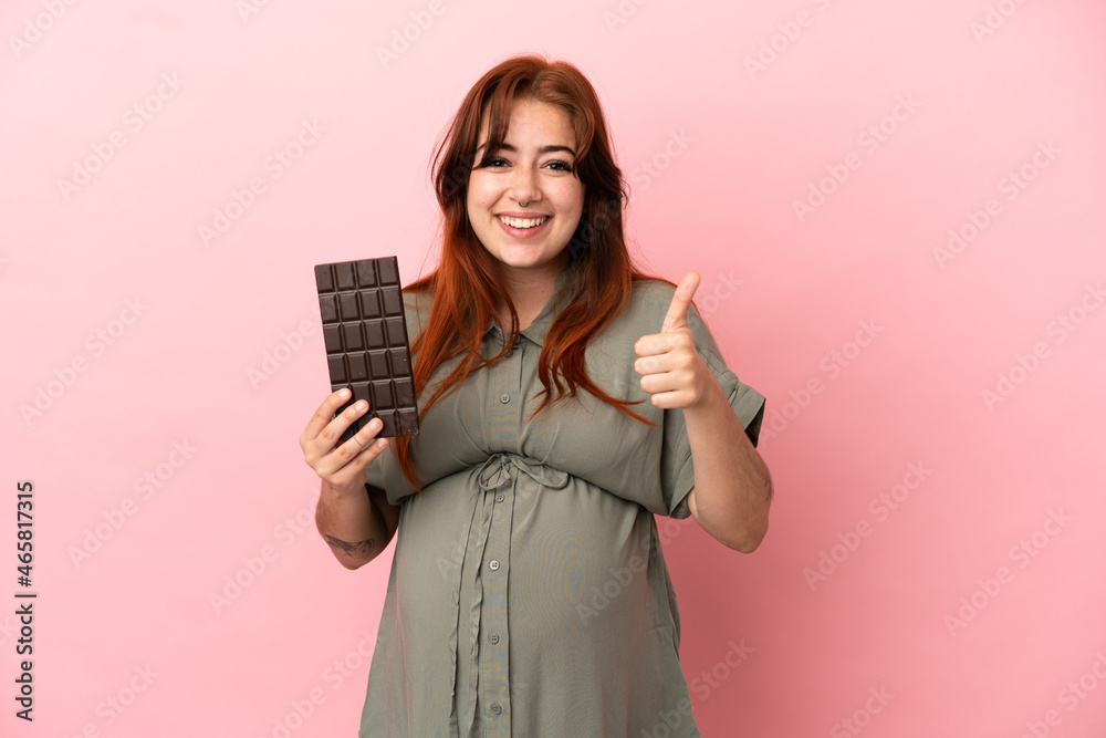 Young redhead caucasian woman isolated on pink background pregnant and holding chocolate with thumb up Young redhead caucasian woman isolated on pink background pregnant and holding chocolate with thumb up