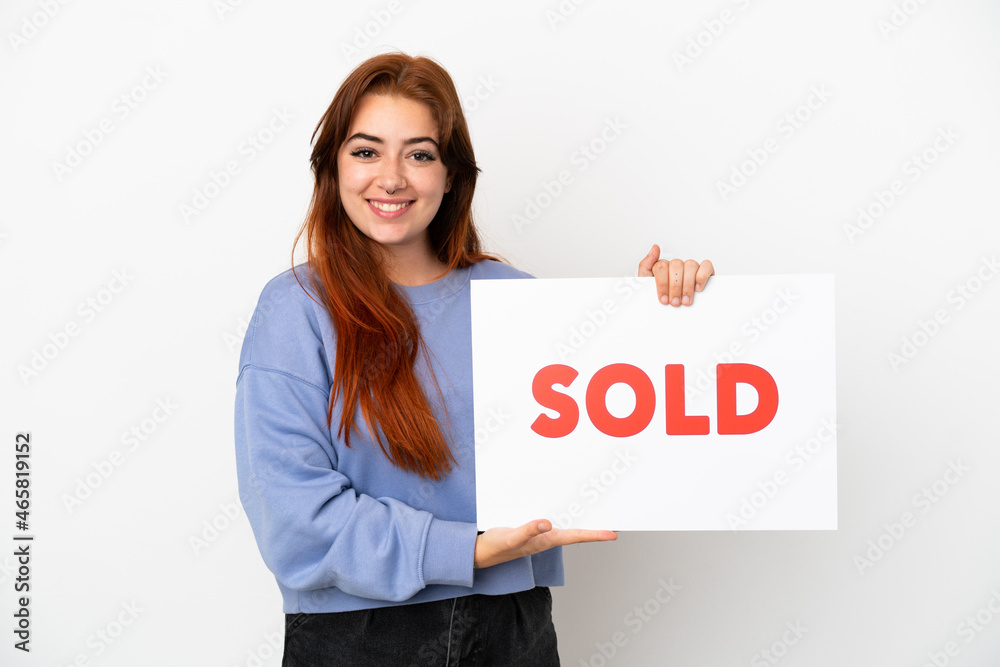 Young redhead woman isolated on white background holding a placard with text SOLD with happy expression Young redhead woman isolated on white background holding a placard with text SOLD with happy expression