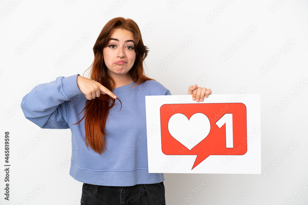 Young redhead woman isolated on white background holding a placard with Like icon and pointing it Young redhead woman isolated on white background holding a placard with Like icon and pointing it