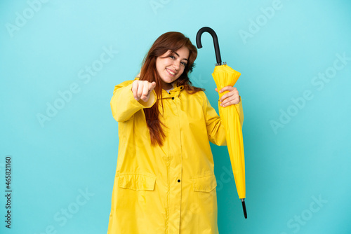 Redhead woman holding an umbrella isolated on blue background pointing front with happy expression
