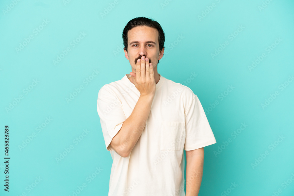 Young caucasian man isolated on blue background covering mouth with hand