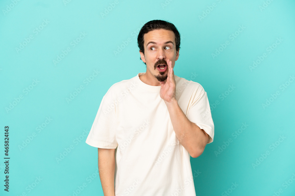 Young caucasian man isolated on blue background whispering something with surprise gesture while looking to the side Young caucasian man isolated on blue background whispering something with surprise gesture while looking to the side