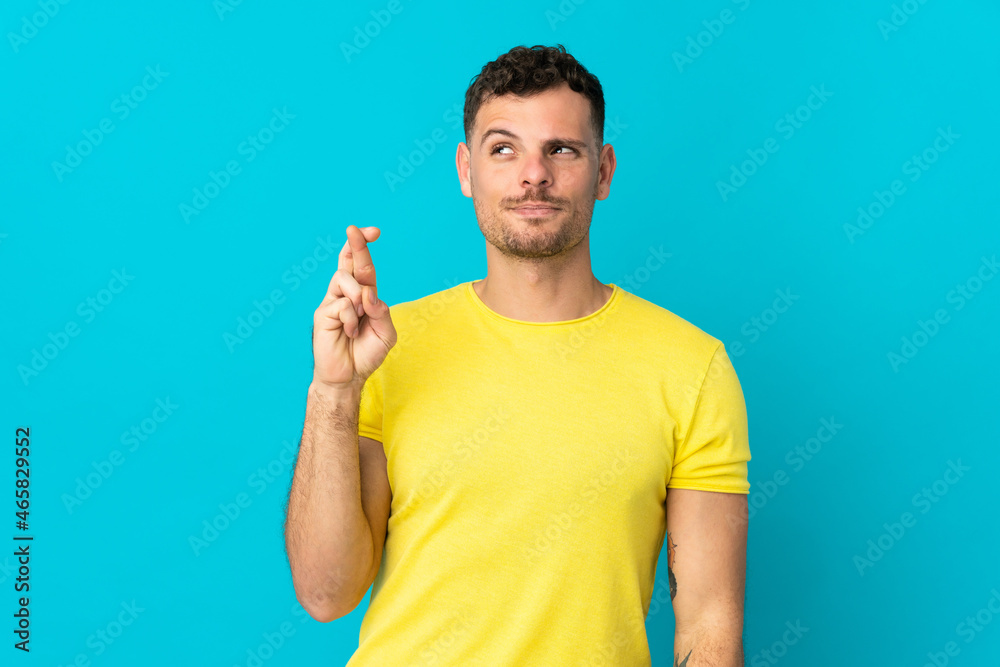 Young caucasian handsome man isolated on blue background with fingers crossing and wishing the best Young caucasian handsome man isolated on blue background with fingers crossing and wishing the best