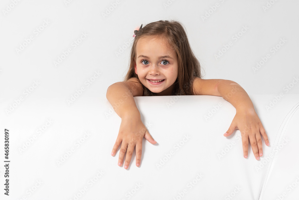 little blonde-haired, sympathetic emotional girl in a colored dress with blond hair leaned on the side of a former leather sofa on a white background, hung up his hands, looked. She is thoughtful little blonde-haired, sympathetic emotional girl in a colored dress with blond hair leaned on the side of a former leather sofa on a white background, hung up his hands, looked. She is thoughtful
